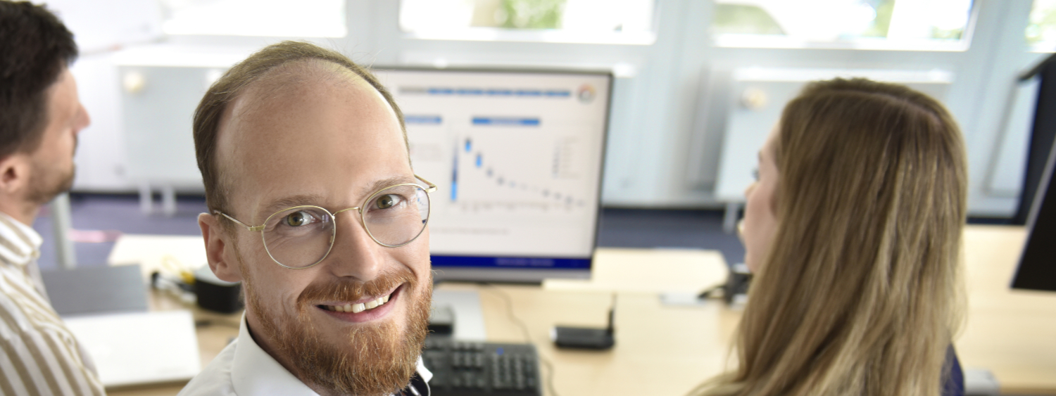Wir bei der apoData schaffen Prozesse, die begeistern. Drei Mitarbeitende der apoData sitzen am Bildschirmarbeitsplatz und einer schaut lächelnd in die Kamera